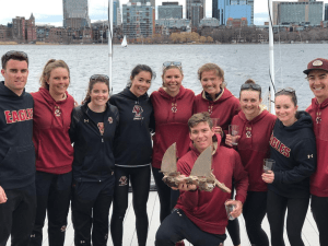 A group of nine people in matching maroon 'EAGLES' athletic gear stand together on a dock, smiling at the camera, with water and a city skyline in the background. One person holds a wooden trophy shaped like boat sails.