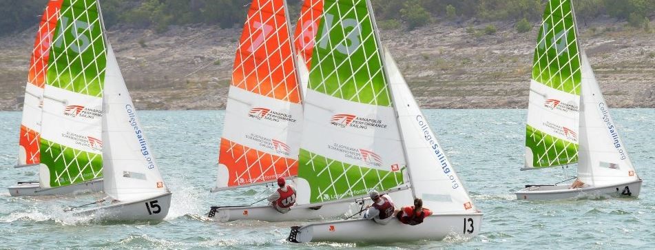 Four small sailboats with colorful orange, green, and white sails race on a lake. Each boat has a team of two sailors wearing life jackets, and the shoreline with trees is visible in the background.