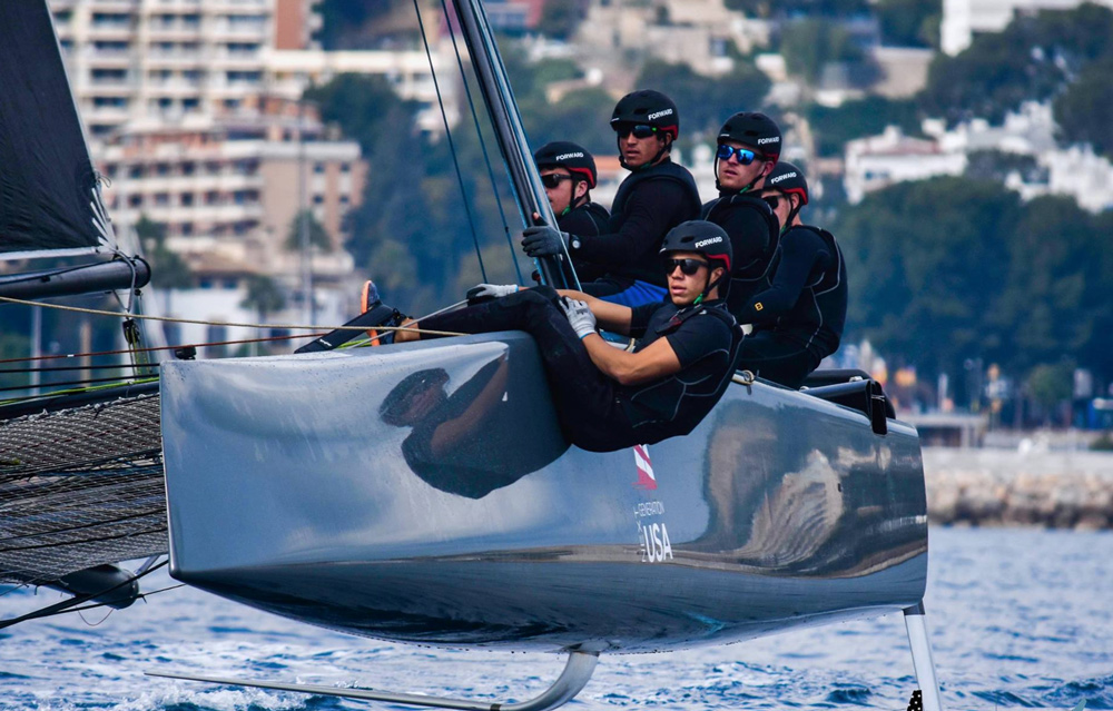 Five sailors wearing helmets and sunglasses lean over the edge of a sleek racing sailboat, focused as they compete on calm blue water with city buildings and trees visible in the background.