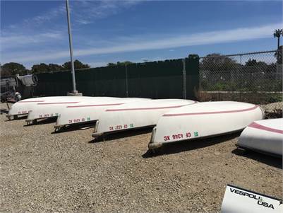 Several small white rowboats with red trim are stored upside down on a gravel lot near a chain-link fence, under a blue sky with scattered clouds.