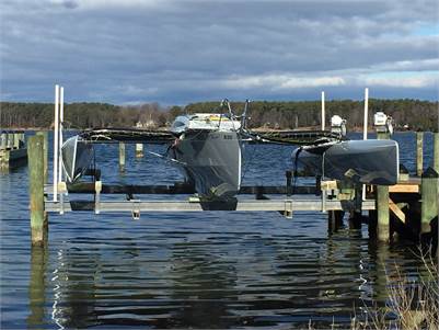 A sleek trimaran sailboat is suspended above the water on a boat lift between two wooden docks, with a forested shoreline and cloudy sky in the background.