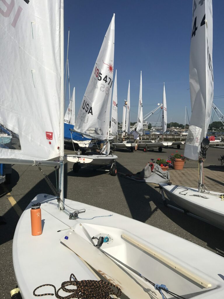 A group of small sailboats with white sails are parked on a paved area near the water under a clear blue sky. An orange water bottle sits on one of the boats in the foreground.