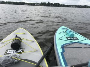 Two paddleboards float side by side on a lake, each with a water bottle and a towel on top. The water is calm, and houses and trees line the distant shore under a cloudy sky.