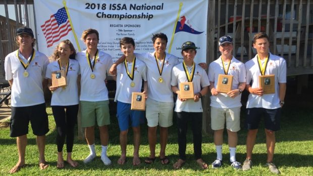 Eight young people wearing medals and holding plaques stand in front of a banner that reads '2018 ISSA National Championship.' They are posing and smiling outdoors on grass near a wooden structure.