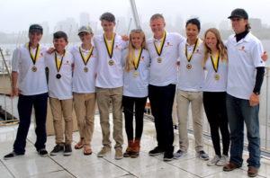 Eight young people and one adult stand side by side, smiling and wearing white polo shirts and medals around their necks, with boats and a city skyline in the foggy background.