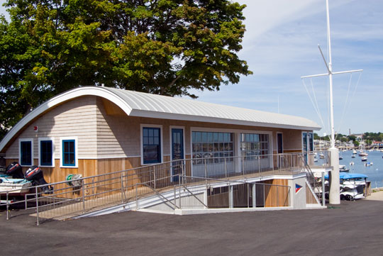 A wooden building with a curved metal roof and blue-trimmed windows sits by a harbor, featuring a ramp for accessibility; boats and water are visible in the background.