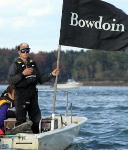 A person in boating gear stands on a small motorboat holding a black flag that reads "Bowdoin." Another person sits nearby. Water and trees are visible in the background.