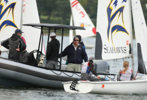 Several people are on boats during a sailing event. Two individuals sit in a small sailboat labeled "Seahawks," while three others are in a larger motorboat nearby, talking. Other sailboats and trees are visible in the background.