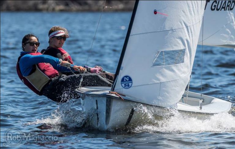 Two people wearing life jackets are leaning out of a small sailboat as it moves quickly through the water, with sunlight reflecting off the waves and the sail marked 'GUARD' partially visible.