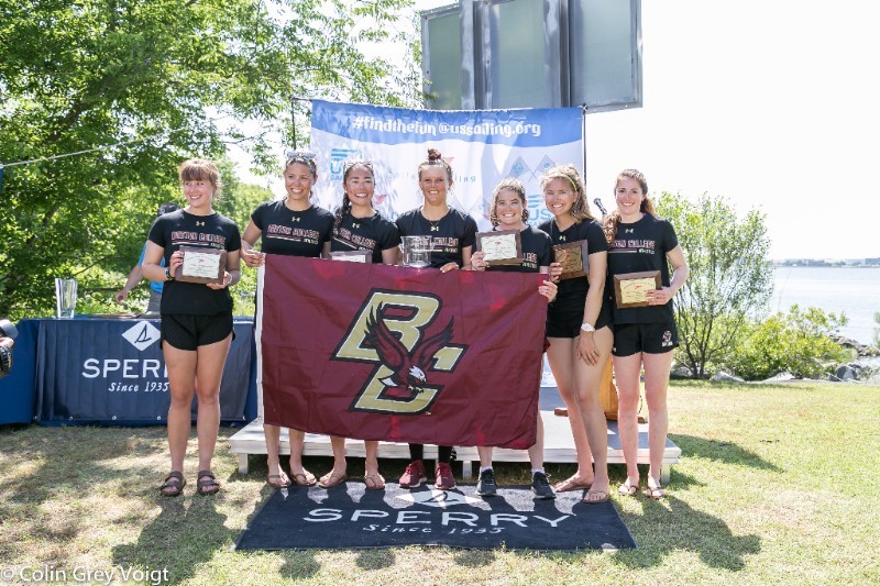 Seven young women in matching athletic outfits hold plaques and a maroon flag with a “BC” logo, posing and smiling outdoors on grass by a lake, in front of a blue Sperry banner and #findthefun usasailing.org sign.