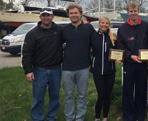 Four people stand outside on grass near boats and cars. Two on the right hold wooden plaques and smile. All are dressed in casual outdoor clothing, and the background includes trees and overcast skies.