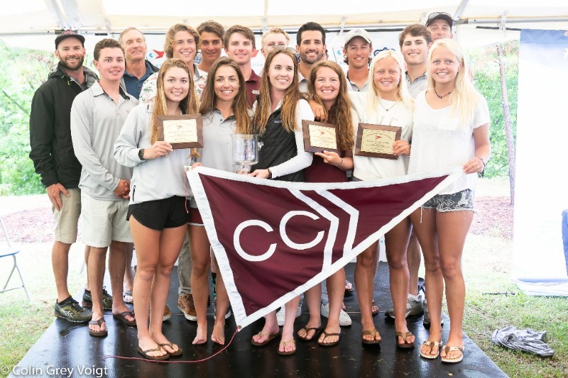 A group of smiling young adults pose together indoors, holding plaques and a maroon flag with "CC" on it, celebrating as a team. Some are wearing medals and casual athletic clothing.