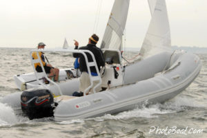 A person in a yellow life jacket sits on a small sailboat near a gray inflatable motorboat with two people. One person on the motorboat gestures toward the sailboat. The boats are on choppy water.