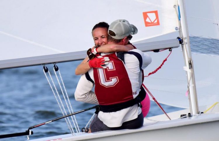 Two sailors on a small sailboat embrace and smile. One wears a red life vest with a large white 'C' on the back and a cap. The water and sail are visible in the background.