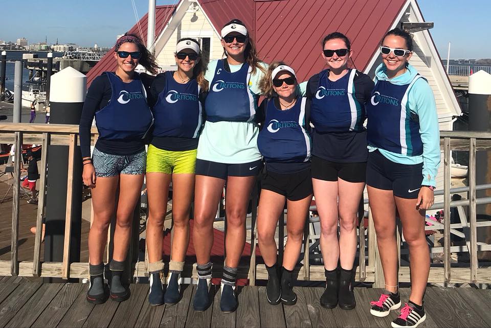 Six women wearing matching navy blue rowing vests and sunglasses stand side by side on a wooden dock, smiling in front of a marina with boats and a building with a red roof.