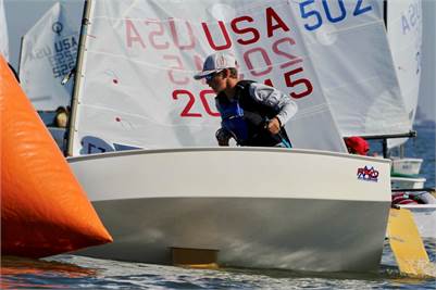A young sailor wearing sunglasses and a cap maneuvers a small sailboat with “USA” and numbers on the sail near a bright orange buoy during a sailing race. Other sailboats are visible in the background.