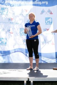 A smiling woman in athletic wear stands barefoot on a stage, holding a large glass trophy. Behind her is a backdrop with sailing logos and text promoting US Sailing and college sailing.