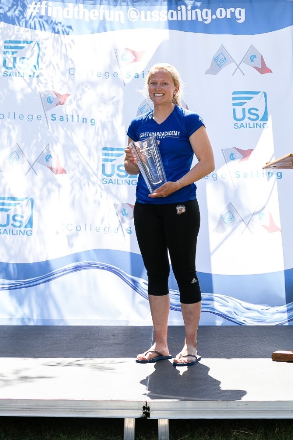 A smiling woman in athletic wear stands barefoot on a stage, holding a large glass trophy. Behind her is a backdrop with sailing logos and text promoting US Sailing and college sailing.
