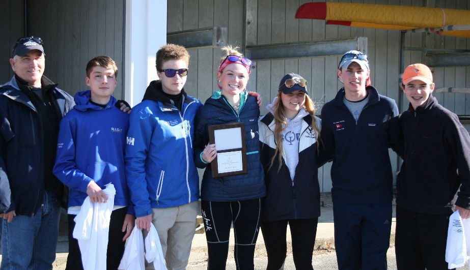 A group of seven people, some holding shirts and one holding a plaque, stand together outside a boathouse, smiling at the camera. A yellow rowing shell is visible in the background.