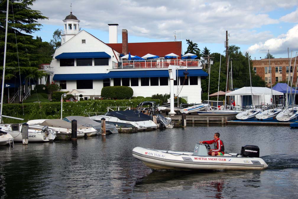 A man drives a small motorboat labeled 'Seattle Yacht Club' on calm water, with a large white yacht club building, docked sailboats, and trees in the background under a partly cloudy sky.