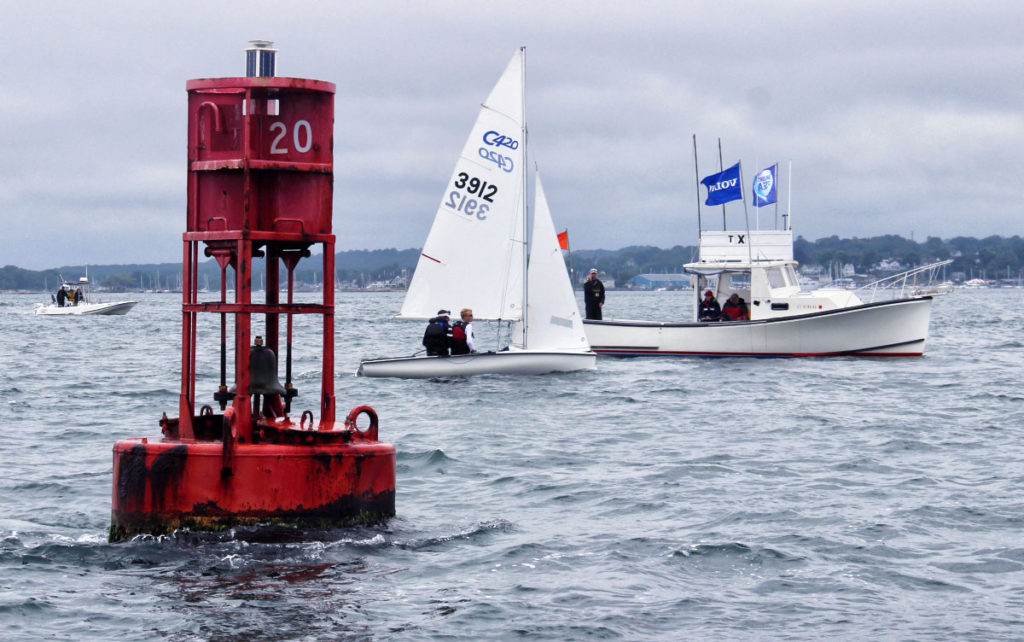 A red buoy marked '20' floats in the foreground, with sailboats and a motorboat on the water nearby. People are visible on the boats, and the shoreline is in the background under a cloudy sky.