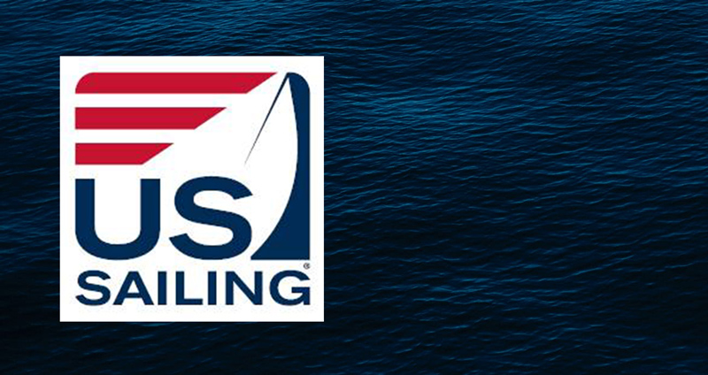 US Sailing logo featuring stylized red stripes and a blue sail, with the words 'US Sailing' below, set against a background of dark blue rippling water.