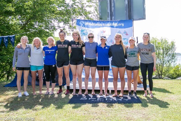 Ten young women stand side by side outdoors, smiling in front of a banner that says US Sailing. They are casually dressed in shorts and t-shirts, with trees and water visible in the background.