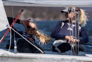 Two young women sailing a boat smile and laugh together. Both wear navy shirts with a white “Y” logo, sunglasses, and visors. The sun shines as they enjoy a breezy day on the water.