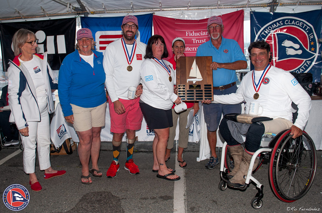 A group of seven people, some wearing medals and sailing attire, pose under a tent. One person in a wheelchair holds a plaque with a sailboat. Various banners hang behind them at an outdoor event.