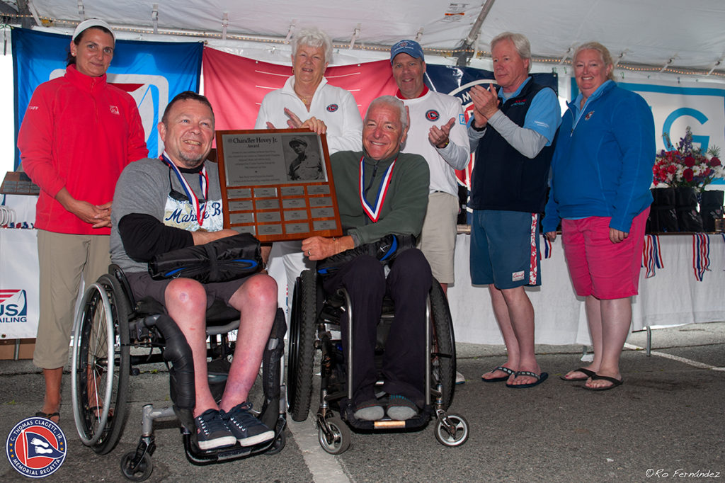 Two men in wheelchairs, smiling and holding a large award plaque, are surrounded by five people clapping and smiling under a tent at a sailing event. Everyone is wearing medals and casual clothes.