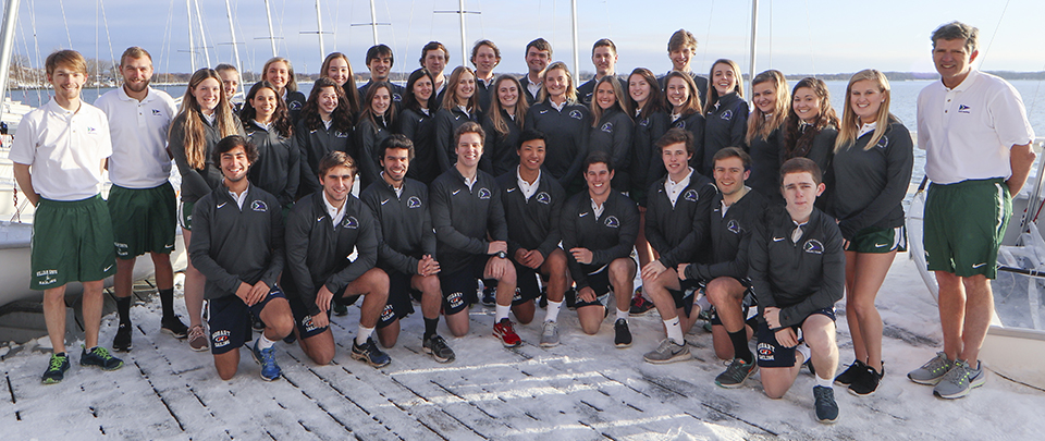 A group of young adults and two coaches posing for a team photo on a dock, with sailboats and water in the background. Most are wearing matching jackets and shorts, smiling at the camera.