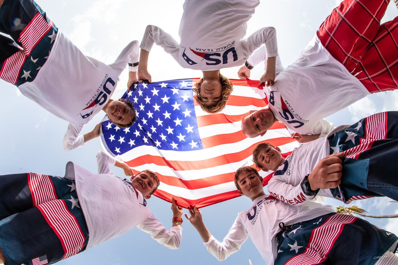 Six people in matching USA shirts and patriotic shorts stand in a circle, holding an American flag above them and looking down at the camera. The flag and their clothing feature red, white, and blue stars and stripes.