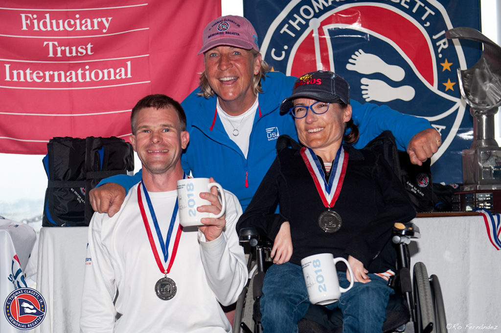 Three smiling people with medals and mugs pose together at an event. A man stands on the left, a woman in a wheelchair is on the right, and another person stands behind them. Event banners are displayed in the background.