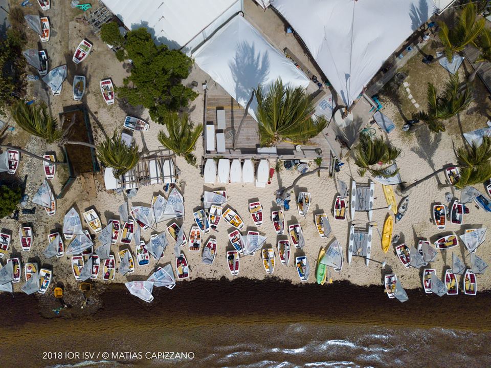 Aerial view of numerous small sailboats and kayaks arranged on a sandy shore near white tents and palm trees, with brown water at the bottom of the image. People are scattered throughout the beach area.