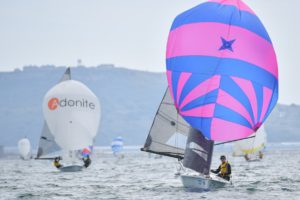 A sailboat with a vibrant pink and blue spinnaker sails on the water, with several other boats in the background, one displaying a white sail labeled "Adonite." Hills and a cloudy sky are visible in the distance.
