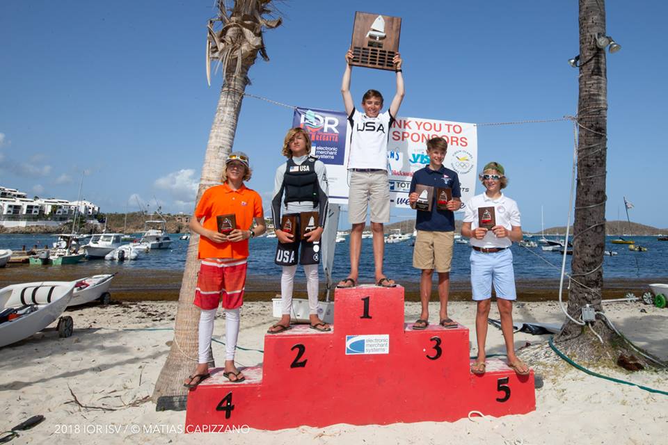 Five young sailors stand on a red winners’ podium on a sandy beach, holding trophies. The first-place winner stands in the center, raising a large trophy above his head. Sailboats, palm trees, and the ocean are in the background.