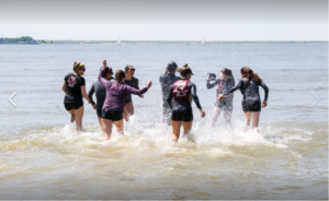 A group of young women wearing matching athletic shirts and shorts splash and play in shallow water at the edge of a lake or sea on a sunny day. Sailboats are visible in the background.