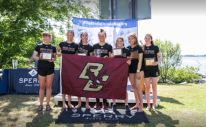 Seven young women in rowing uniforms stand outdoors holding plaques and a Boston College flag, smiling for a team photo. They are in front of a table and banners with "Sperry" and #findtheinussailing.org displayed.