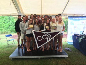 A group of young adults stand together under a tent, smiling and holding plaques and a maroon and white flag with "CC" on it, at what appears to be a sailing or collegiate event.