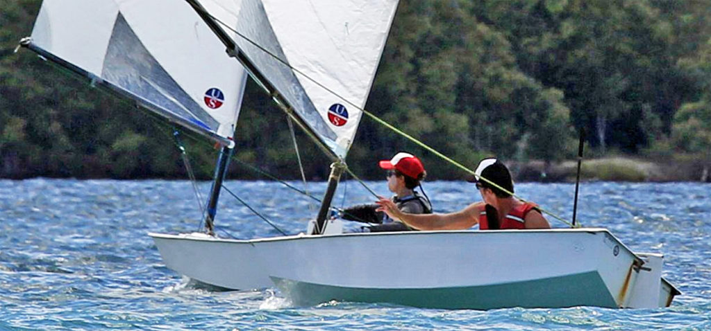 Two people wearing life jackets and hats are sailing small white boats on a blue body of water, surrounded by green trees in the background. The scene appears sunny and calm.