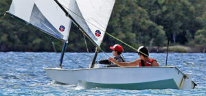 Two people wearing life jackets and hats are sailing small white boats on a blue body of water, surrounded by green trees in the background. The scene appears sunny and calm.