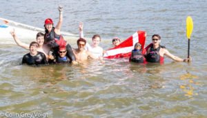 A group of people in wetsuits and swimwear celebrate in a lake, some raising their arms and holding an MIT flag and a paddle. They appear joyful, with a boat partially visible in the background.