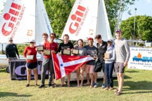 A group of eight people stand in front of sailboats, holding plaques and a red and white nautical flag, smiling at the camera during a sunny awards event.