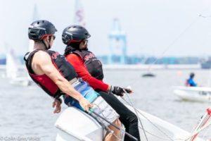 Two people wearing helmets and life vests lean out from a small sailboat on the water, balancing as they navigate. Other sailboats and a port with cranes are visible in the background.