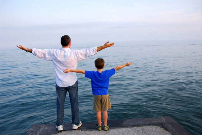 A man and a young boy stand on a pier facing the ocean with their arms outstretched, mimicking each other, under a clear sky.