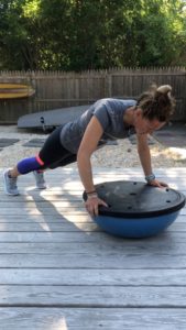 A woman in athletic clothing does a push-up with her hands on an upside-down BOSU ball on a wooden deck outside, with a wooden fence and greenery in the background.