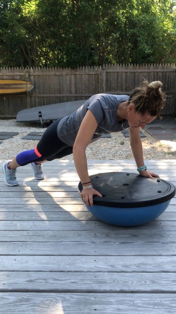 A woman in athletic clothing does a push-up with her hands on an upside-down BOSU ball on a wooden deck outside, with a wooden fence and greenery in the background.