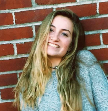 A young woman with long blonde hair, wearing a grey sweater and a black choker necklace, smiles while leaning against a red brick wall.