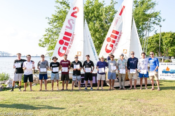 A group of people stand in a row on grass by the water, holding certificates. Sailboats with tall sails and event banners are behind them. Trees and a bridge are visible in the background on a sunny day.