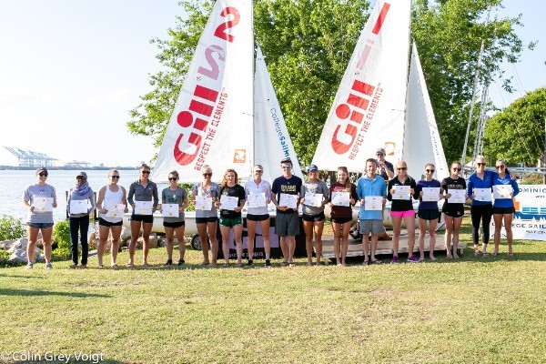 A group of people stand in a row outdoors on grass, holding certificates in front of three sailboats with "GILL" on the sails. Water and trees are visible in the background on a sunny day.
